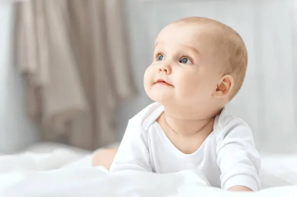En bebis i vit body ligger på mage i en säng, lyfter huvudet och tittar uppåt