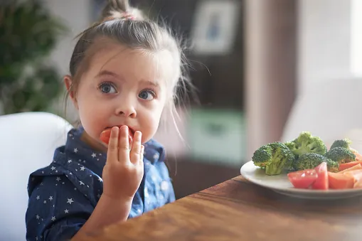 En liten flicka vid bordet äter med fingrarna, med en tallrik broccoli, tomat och morot framför sig