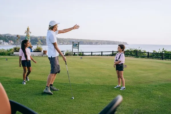 En vuxen med golfklubba visar något för ett barn på en golfbana vid havet, två barn står bredvid