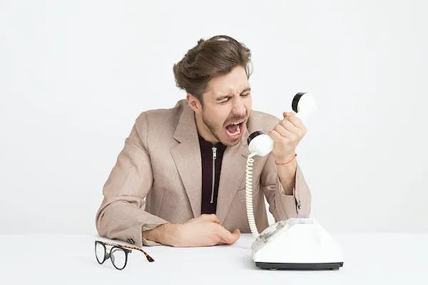 En man i beige kavaj skriker i luren på en gammal vit snurrtelefon, glasögonen ligger på bordet