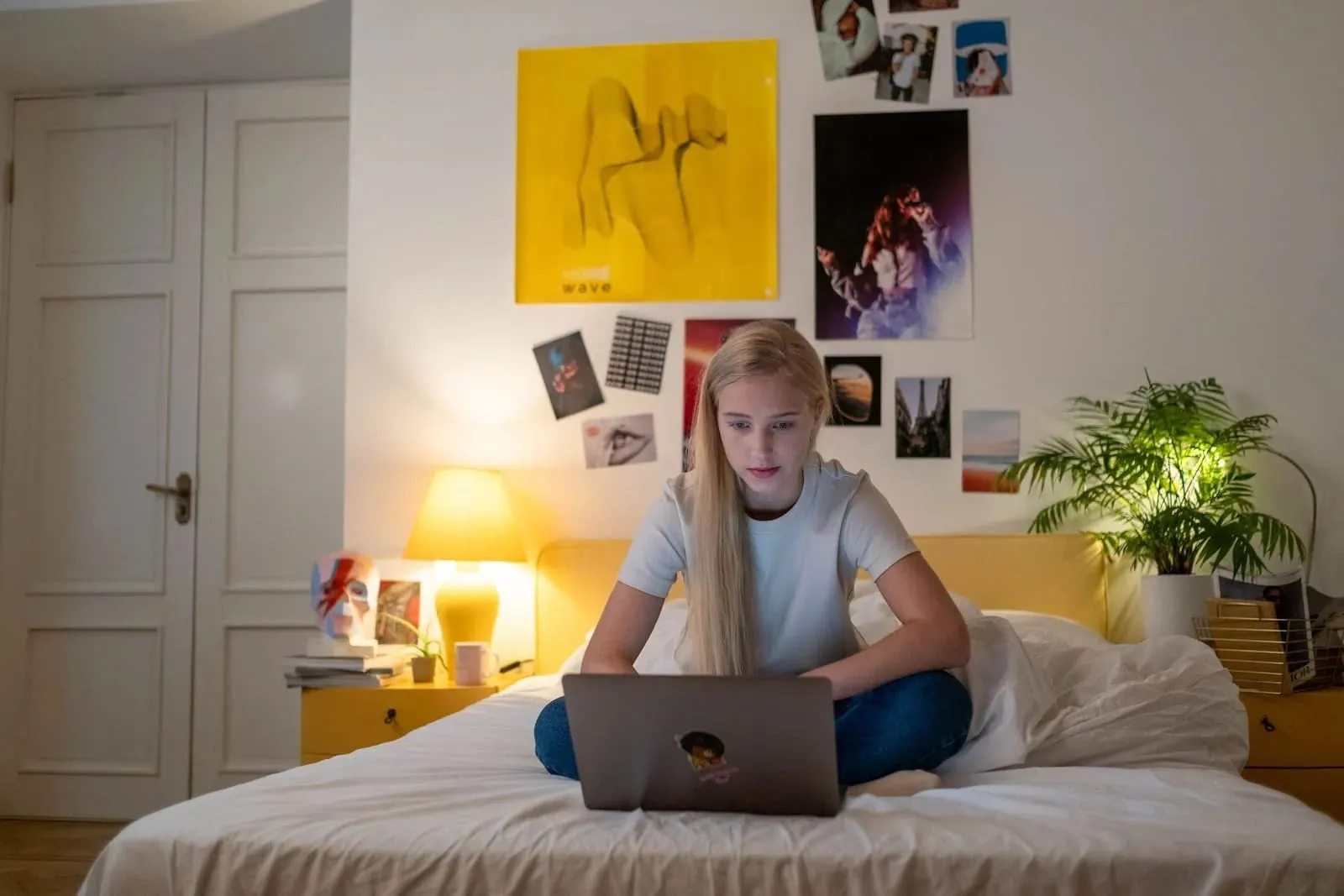En tonårsflicka sitter med benen i kors på sängen med sin laptop i ett rum fullt av posters och foton
