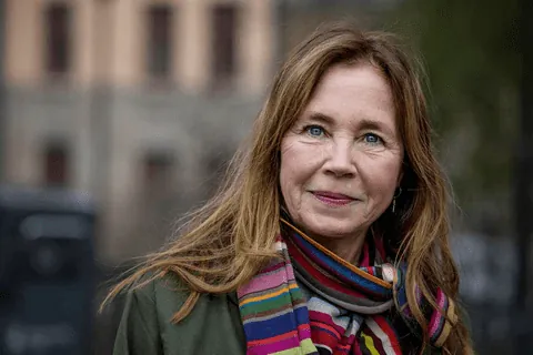 Kvinna med långt rödbrunt hår i grön jacka och färgglad randig halsduk, utomhus framför en ljus byggnad